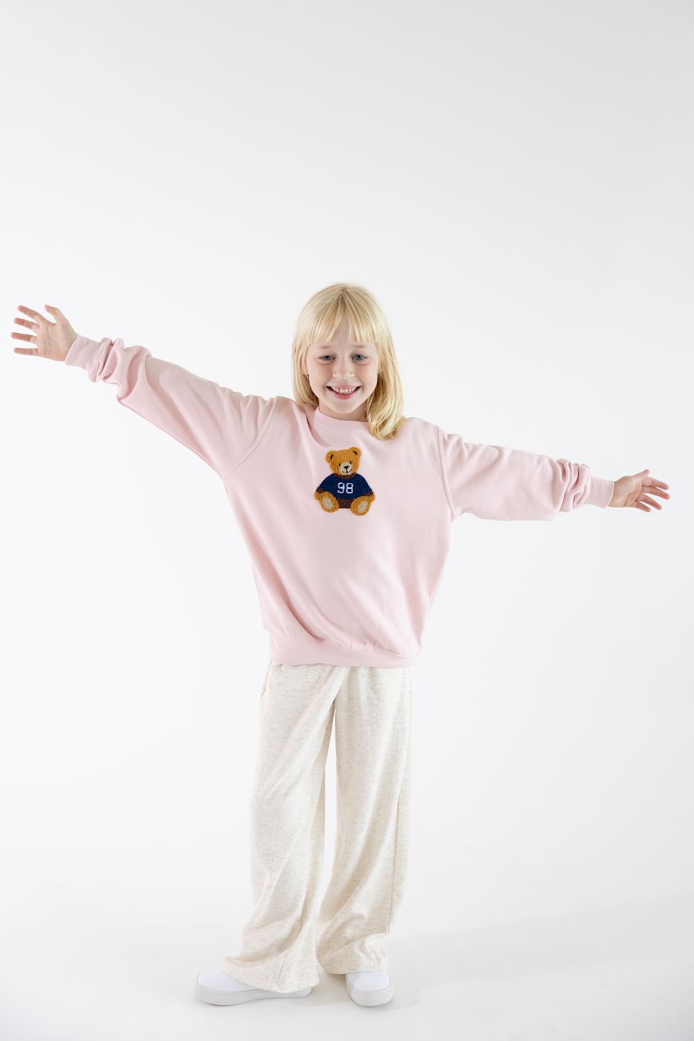 Young girl with blonde hair smiling with arms outstretched, wearing a light pink crewneck sweatshirt with a teddy bear patch and light-colored pants.