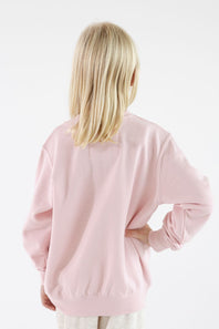 Back profile of a young girl with blonde hair wearing a light pink long-sleeve crewneck sweatshirt with ribbed cuffs and hem.