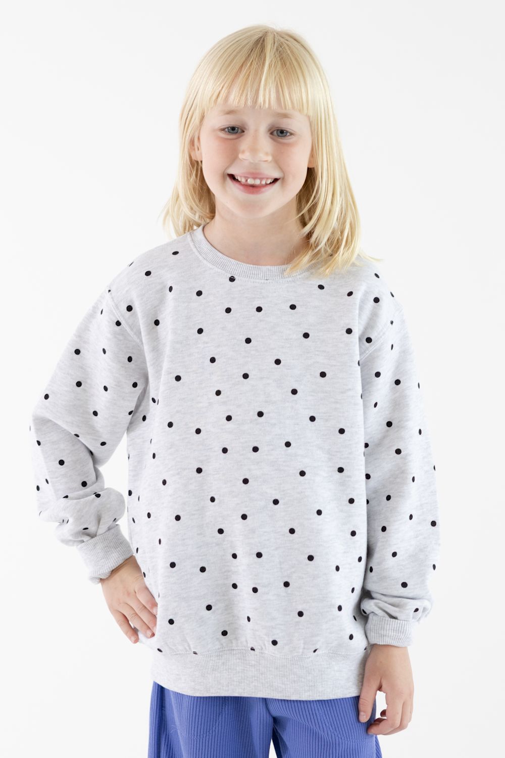 A young blonde girl smiling while wearing a light gray long-sleeve sweatshirt with a black polka dot pattern, paired with blue pants, against a plain white background.