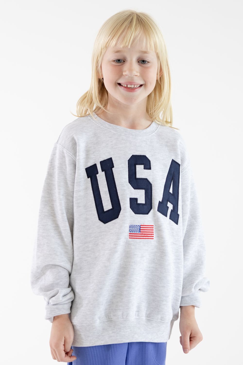 A young blonde girl smiling while wearing a light gray heather crewneck sweatshirt with 'USA' printed in navy blue letters and a small American flag graphic below, paired with blue pants against a white studio background.