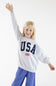 A young blonde girl smiling and waving with one arm raised, wearing a light gray sweatshirt with 'USA' printed in navy blue letters and a small American flag graphic, paired with blue pants, against a plain white studio background.