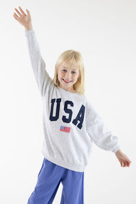 A young blonde girl smiling and waving with one arm raised, wearing a light gray sweatshirt with 'USA' printed in navy blue letters and a small American flag graphic, paired with blue pants, against a plain white studio background.