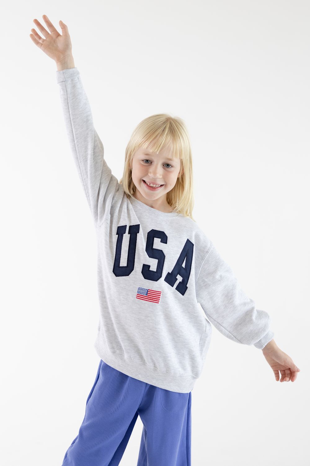 A young blonde girl smiling and waving with one arm raised, wearing a light gray sweatshirt with 'USA' printed in navy blue letters and a small American flag graphic, paired with blue pants, against a plain white studio background.