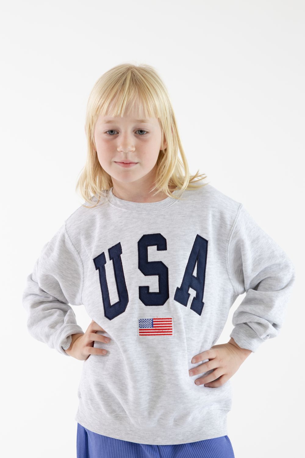 A young blonde girl wearing a light gray heather sweatshirt with 'USA' in navy blue letters and a small American flag graphic, paired with blue pants, against a plain white background.