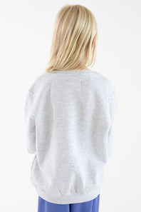 A blonde girl viewed from behind wearing a light gray heather crewneck sweatshirt and blue pants, set against a plain white studio background.