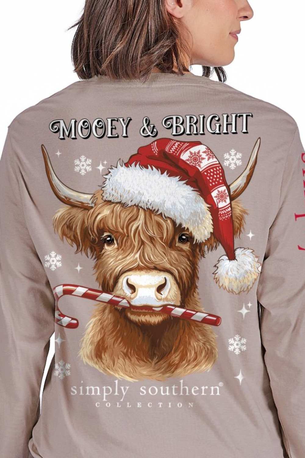 A woman wearing a taupe long-sleeve shirt, viewed from the back. The shirt features a large graphic of a Highland cow wearing a red Santa hat and holding a candy cane, with 'Mooey & Bright' and 'simply southern' collection' printed on the back.