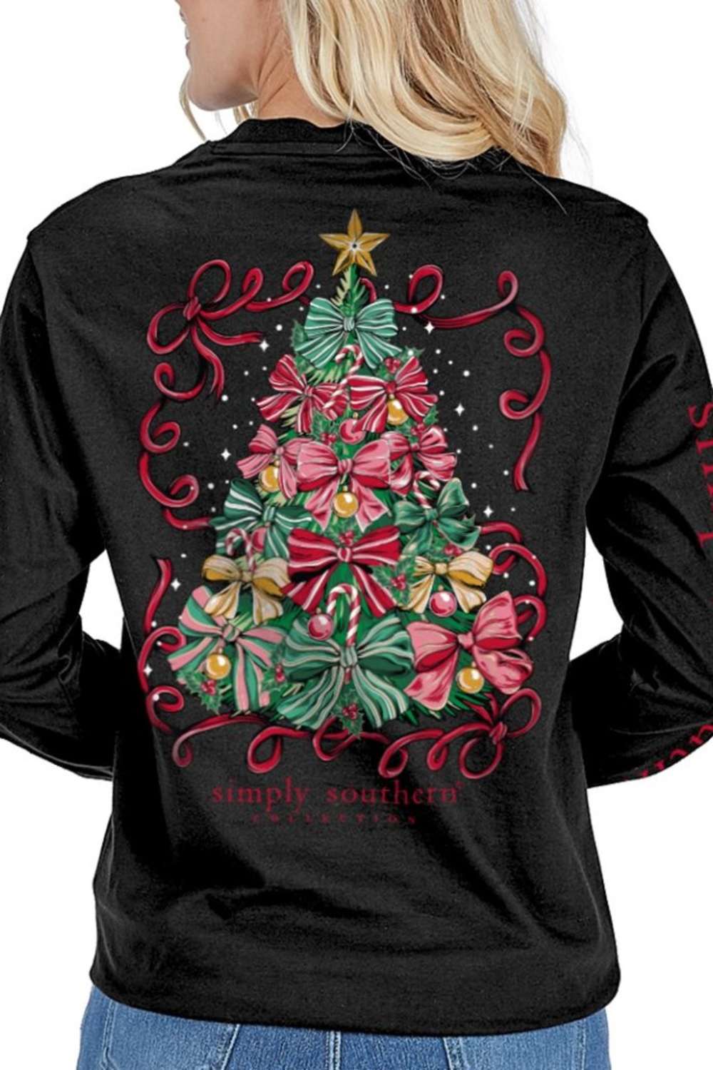A woman wearing a black long-sleeve shirt, viewed from the back. The shirt features a large graphic of a Christmas tree decorated with red and pink bows, green pine needles, a gold star, and red scrollwork, with the text 'simply southern' 
