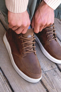 A close-up of a person's hands adjusting the lace of a brown leather sneaker, with the other shoe visible on a weathered wooden deck.