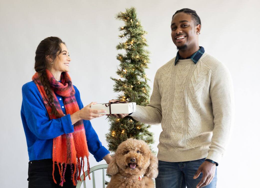 Man and woman holding a gift in front of a decorated Christmas tree with a dog.