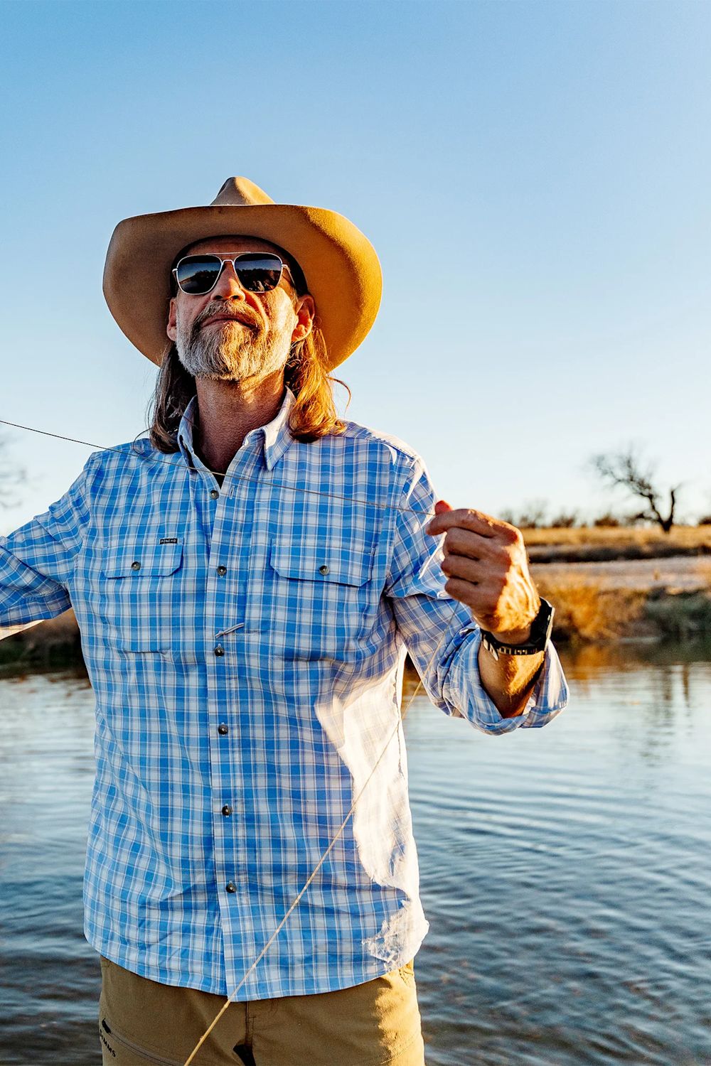A man wearing a tan cowboy hat, blue and white plaid button-down shirt, and sunglasses stands by a river, holding a fishing line. The background shows a calm river, distant trees, and a clear blue sky.