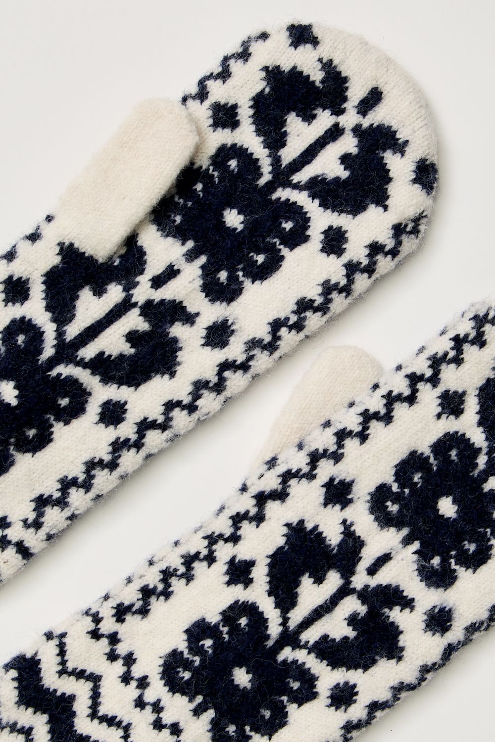 A close-up of a pair of knit mittens with a dark navy blue and off-white Nordic floral pattern, showing the soft fleece lining of one mitten.