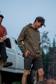 A man wearing an olive green hooded sweatshirt, dark gray shorts, and a black baseball cap stands next to a white pickup truck outdoors. Another man is partially visible on the left.