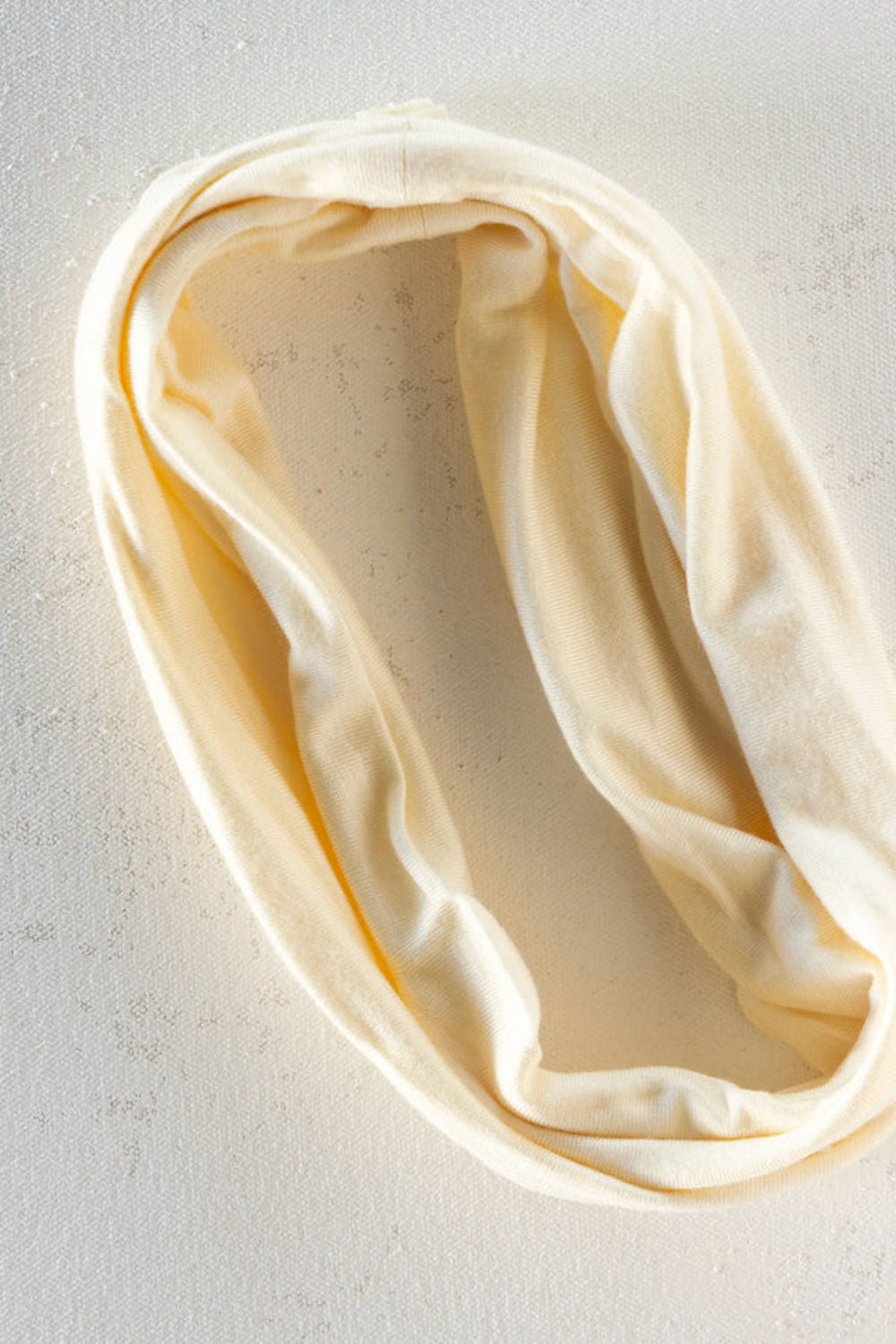 A cream-colored infinity loop neck warmer, draped in soft folds on a textured light gray background.