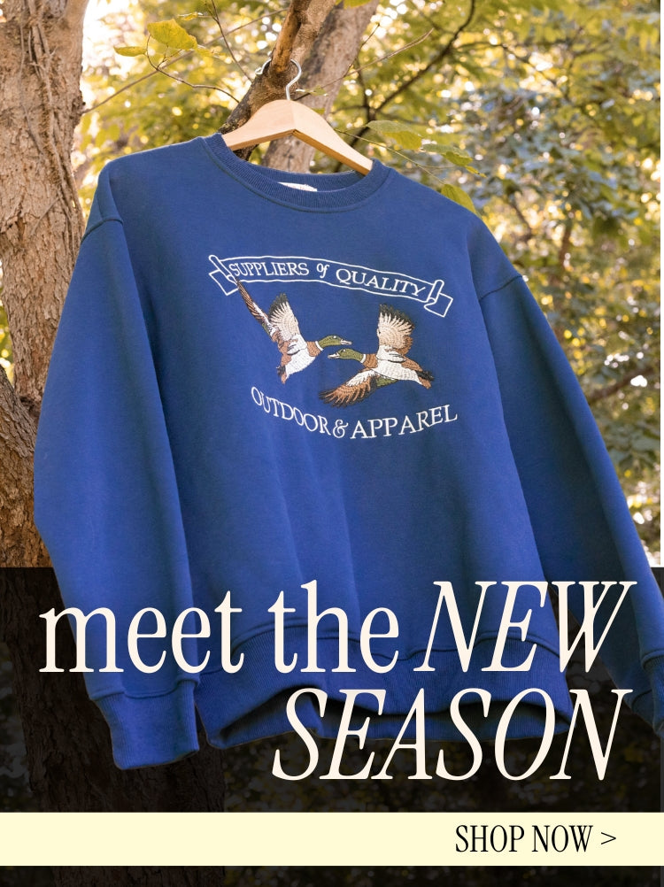 Blue sweatshirt with embroidered birds and text hangs on a tree branch outdoors. Overlay text reads, “meet the NEW SEASON, SHOP NOW.”.