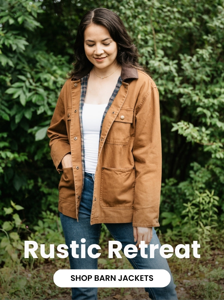 A model wears a brown barn jacket with a plaid lining, white top, and blue jeans against a lush green backdrop. Text reads Rustic Retreat and Shop Barn Jackets