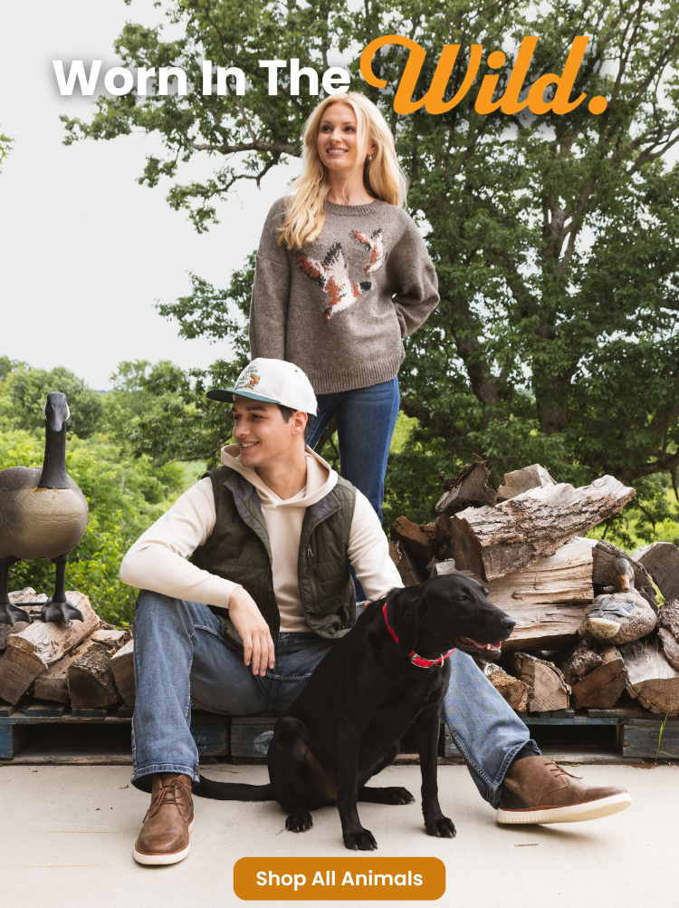 Two models pose outdoors with a black dog in front of stacked firewood, showcasing casual fall apparel. One model stands in a brown wildlife graphic sweater and blue jeans, while the other sits wearing a cream hoodie, olive quilted vest, light wash jeans, brown shoes, and a white cap. The image includes the text "Worn In The Wild." and a "Shop All Animals" call-to-action button.