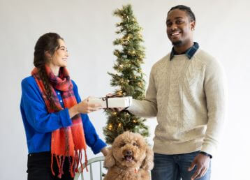 Two people exchanging a gift in front of a decorated Christmas tree with a dog present.