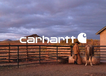 A cowboy wearing a hat, brown jacket, and chaps stands next to a light-colored horse drinking from a water trough inside a fenced enclosure. Rolling hills and a cloudy, twilight sky fill the background. The white Carhartt logo is centered prominently over the scene