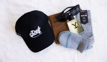 Black cap with 'Dad Gang' text, wallet sunglasses, cologne and socks on a white surface