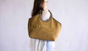 A model wearing a white knit poncho carries a large tan corduroy tote bag over one shoulder, shown from the back against a neutral gray background.