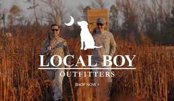 Local Boy Outfitters promotional banner featuring two men in camouflage outdoor clothing standing in a field of tall grasses, with the brand’s dog-and-moon logo and a Shop Now call to action