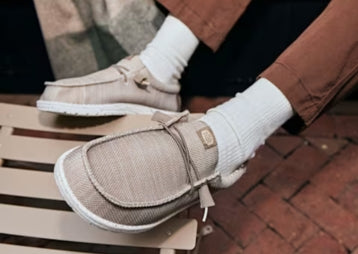 Close-up of beige slip-on shoes with decorative ties and white soles, styled with white socks and brown pants while resting on a wooden bench.
