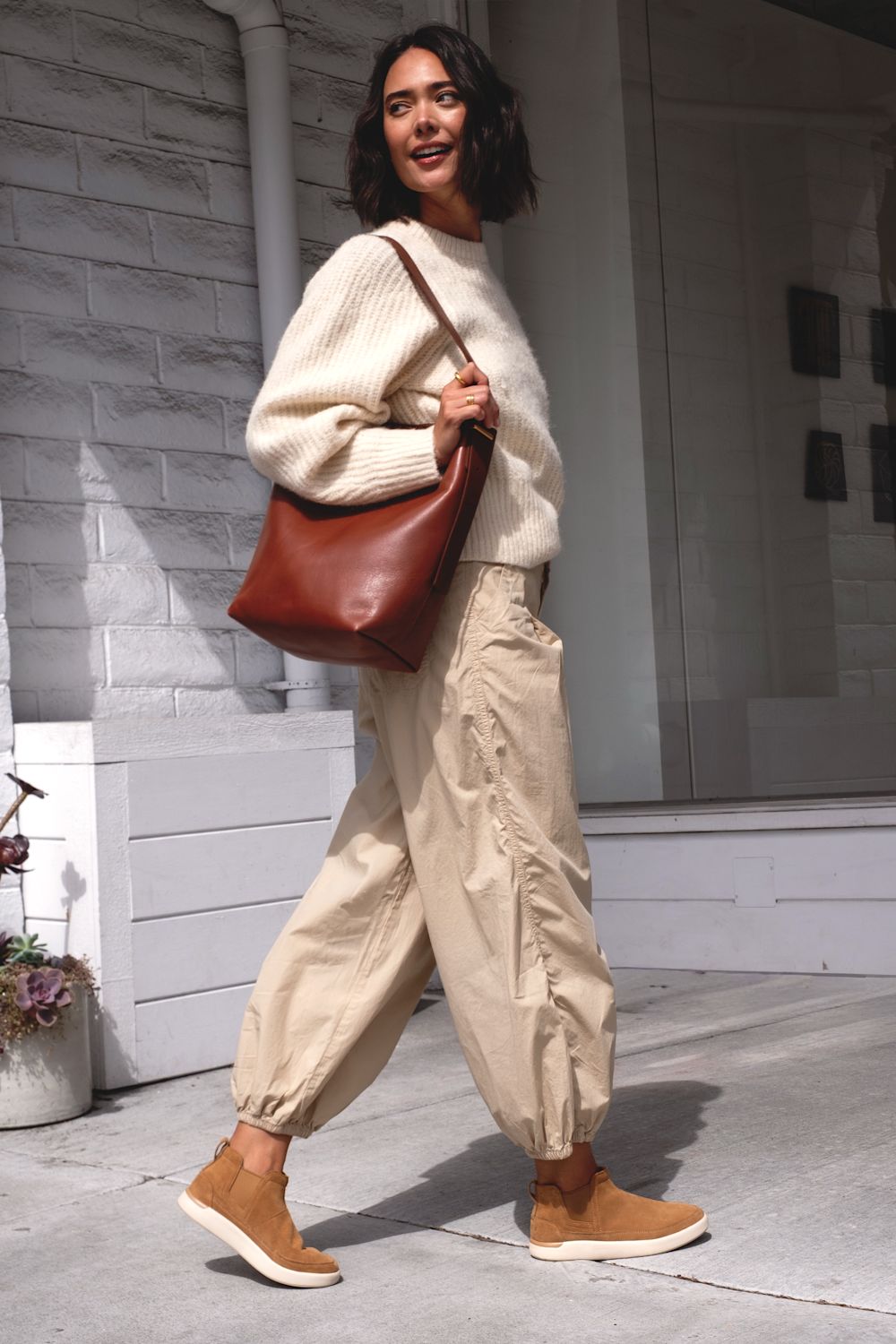 A woman walking outdoors wearing a cream ribbed knit sweater, beige baggy pants, tan suede boots, and carrying a brown leather shoulder bag.