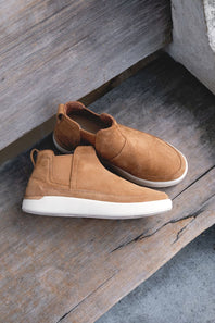 A pair of tan suede slip-on sneakers with elastic side panels and thick white soles, resting on rustic wooden steps.