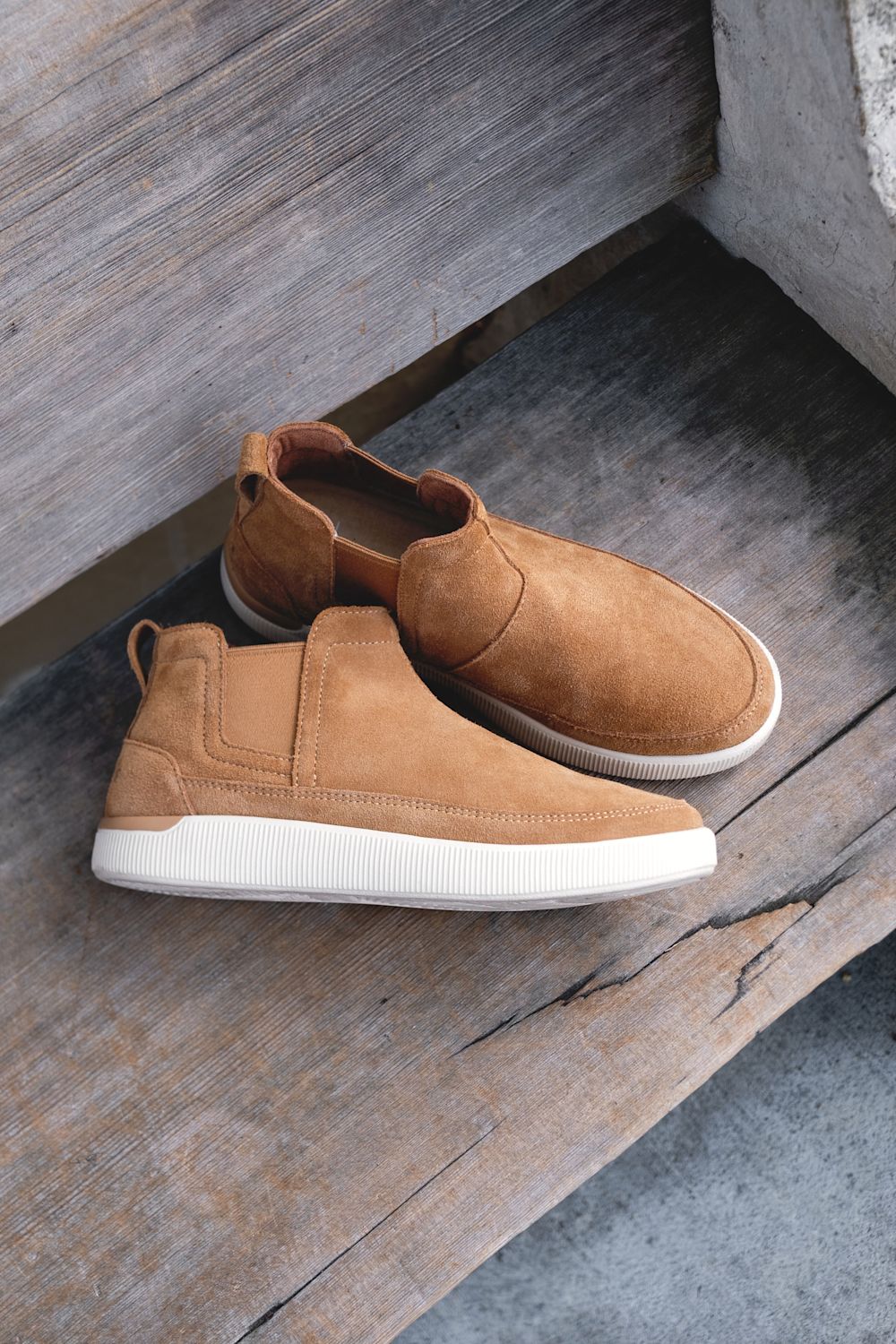 A pair of tan suede slip-on sneakers with elastic side panels and thick white soles, resting on rustic wooden steps.