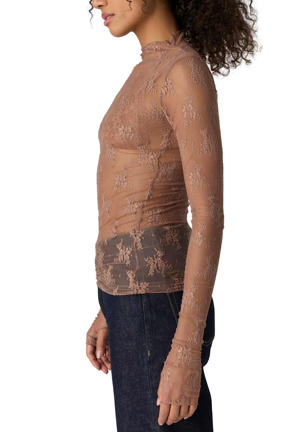 A woman shown in side profile wearing a sheer, long-sleeve mock-neck lace top in a warm taupe-brown color, paired with dark blue jeans, against a plain white background.