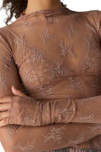 Close-up of a person wearing a sheer taupe-brown long-sleeve top with floral lace applique and a high mock neck.