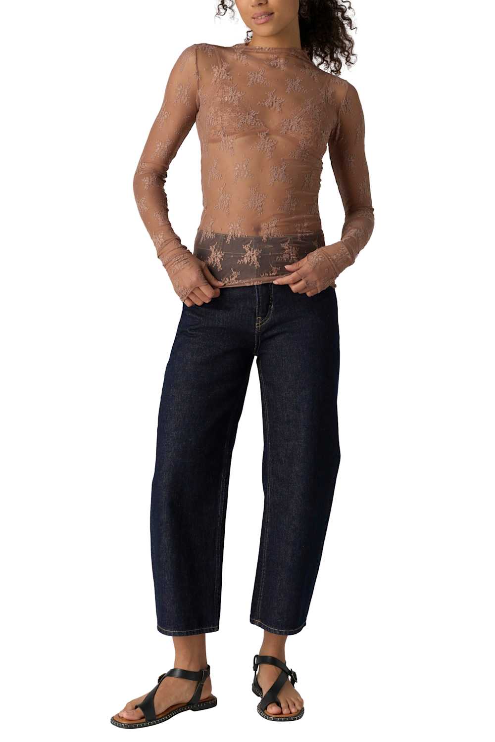 A model wearing a sheer, patterned brown mock-neck long-sleeve top paired with dark blue cropped jeans and black sandals, shown against a plain white background.