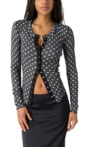 A woman wearing a black long-sleeve cardigan with a white polka-dot pattern and brown buttons, paired with a black skirt, against a plain white background.