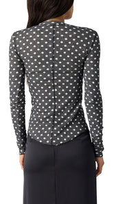 A woman shown from the back wearing a long-sleeve fitted top with a black base and small white floral or dot pattern, paired with a dark skirt against a plain white studio background.