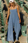 FP Movement Hot Shot Strappy Onesie for Women in Blue Ash; Woman wearing a blue jumpsuit standing in an outdoor setting with green chairs.
