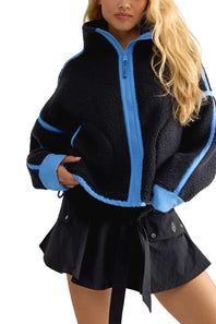 A woman wearing a black sherpa fleece zip-up jacket with bright blue trim and a black pleated mini skirt, photographed against a plain white background.