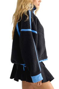 A woman wearing a black sherpa fleece cropped jacket with light blue contrast trim and a matching black pleated mini skirt, shown from a side profile against a white background.