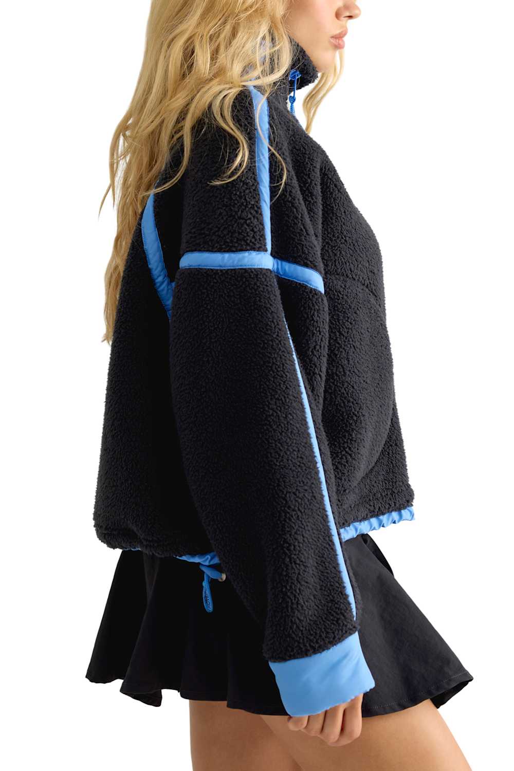 A woman wearing a black sherpa fleece cropped jacket with light blue contrast trim and a matching black pleated mini skirt, shown from a side profile against a white background.