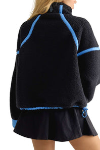 A person shown from the back wearing a black fleece pullover with bright blue contrast trim and a black pleated skirt against a plain white studio background.