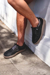 A person wearing black suede low-top sneakers with tan soles, sitting against a white painted brick wall outdoors.