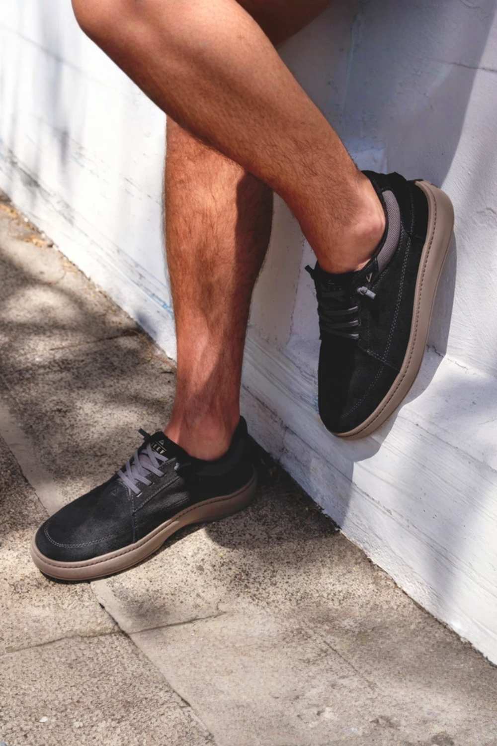 A person wearing black suede low-top sneakers with tan soles, sitting against a white painted brick wall outdoors.