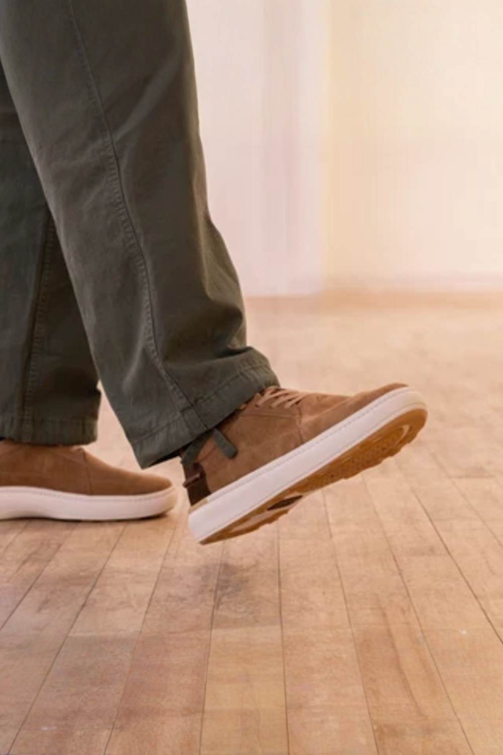 A person wearing brown suede sneakers with white soles and olive green pants, captured mid-step on a light wood plank floor.