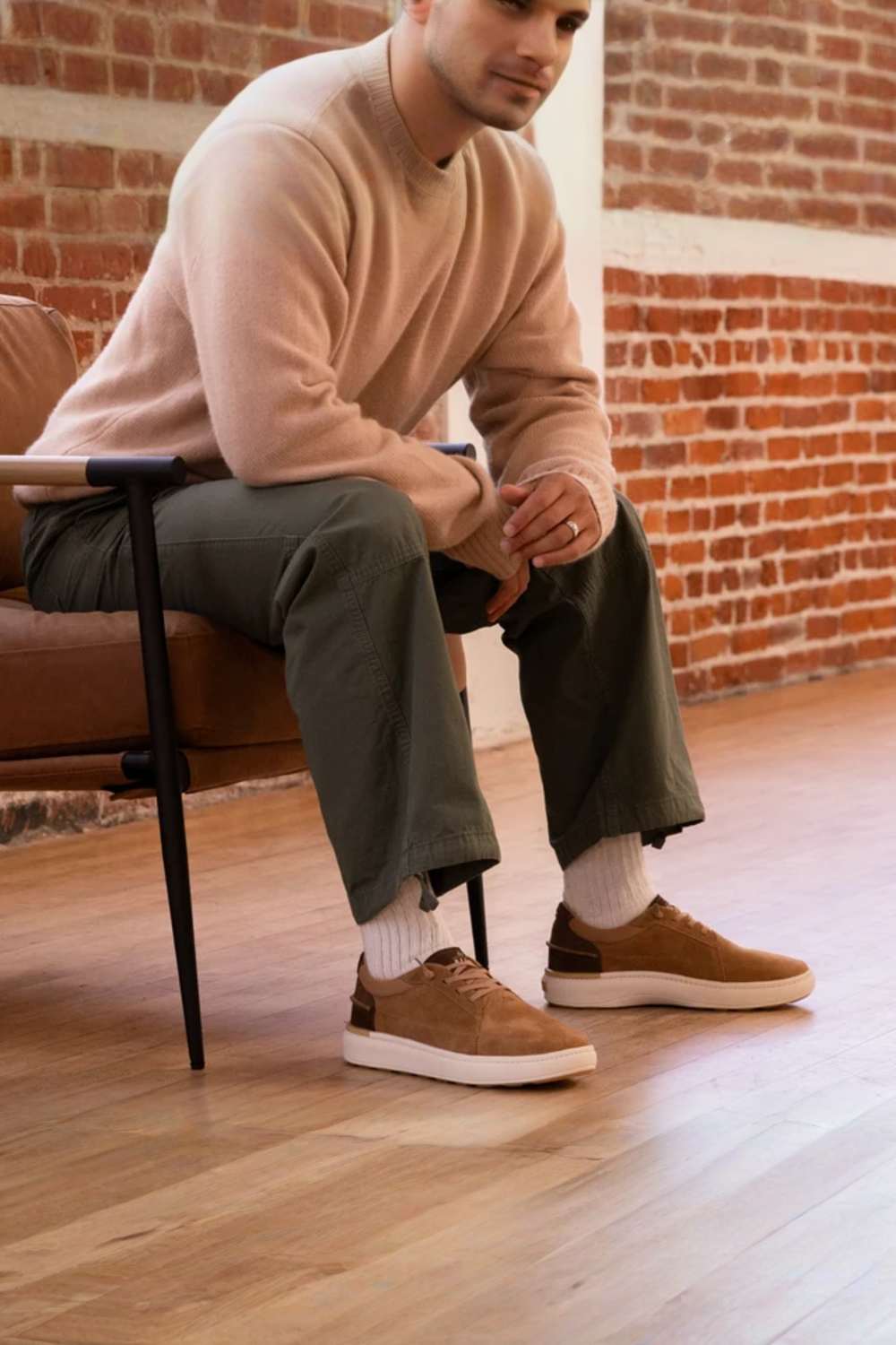 A man sitting on a brown leather chair in a room with a brick wall and wood floor, wearing a light beige knit sweater, olive green pants, white socks, and brown suede sneakers.