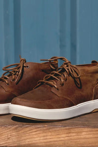 A pair of brown suede low-top sneakers with white soles, displayed on a rustic wooden surface against a blue wooden background.