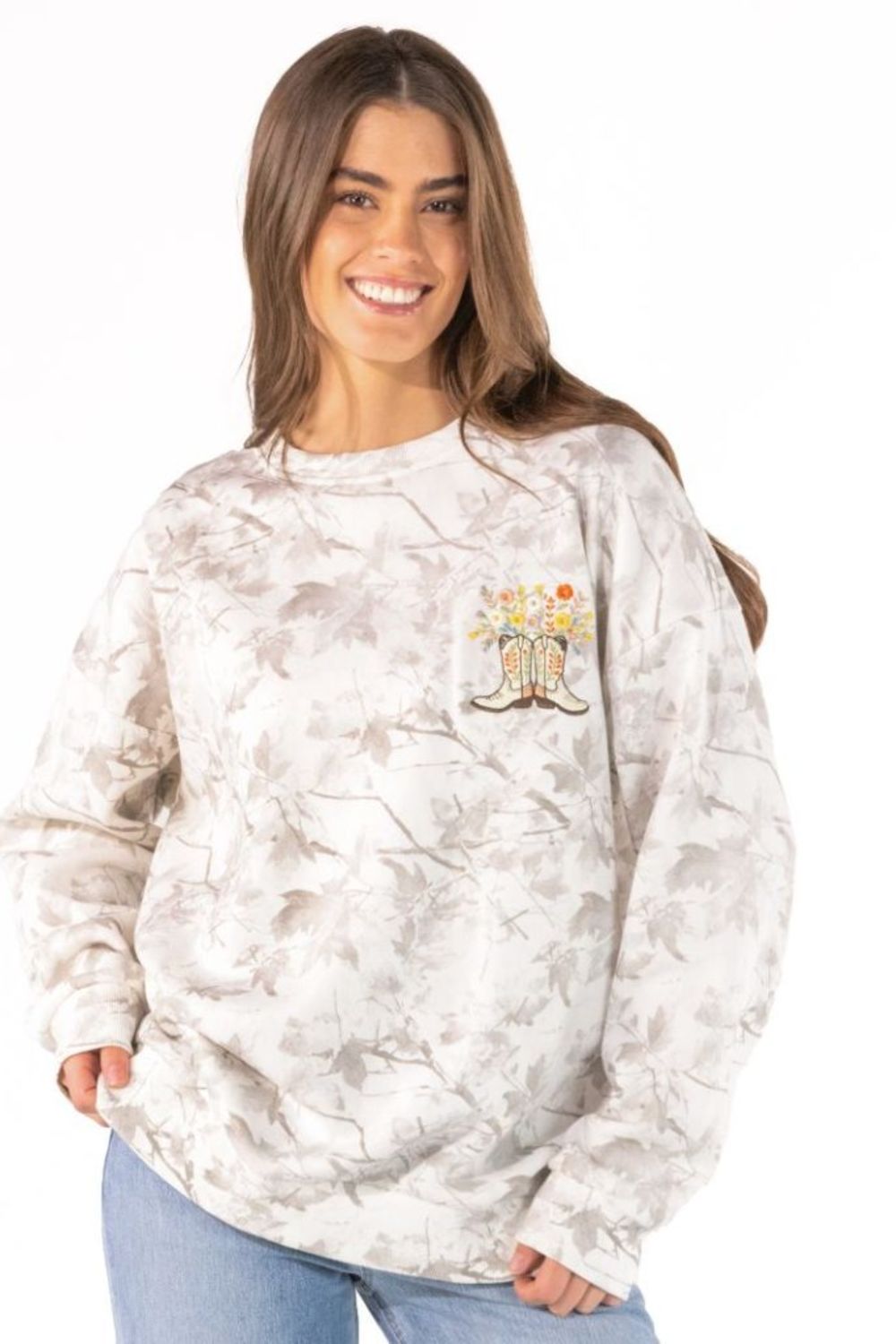 A woman wearing a light beige and white tie-dye sweatshirt with a small graphic of cowboy boots and flowers on the left chest, paired with blue jeans, against a plain white background.