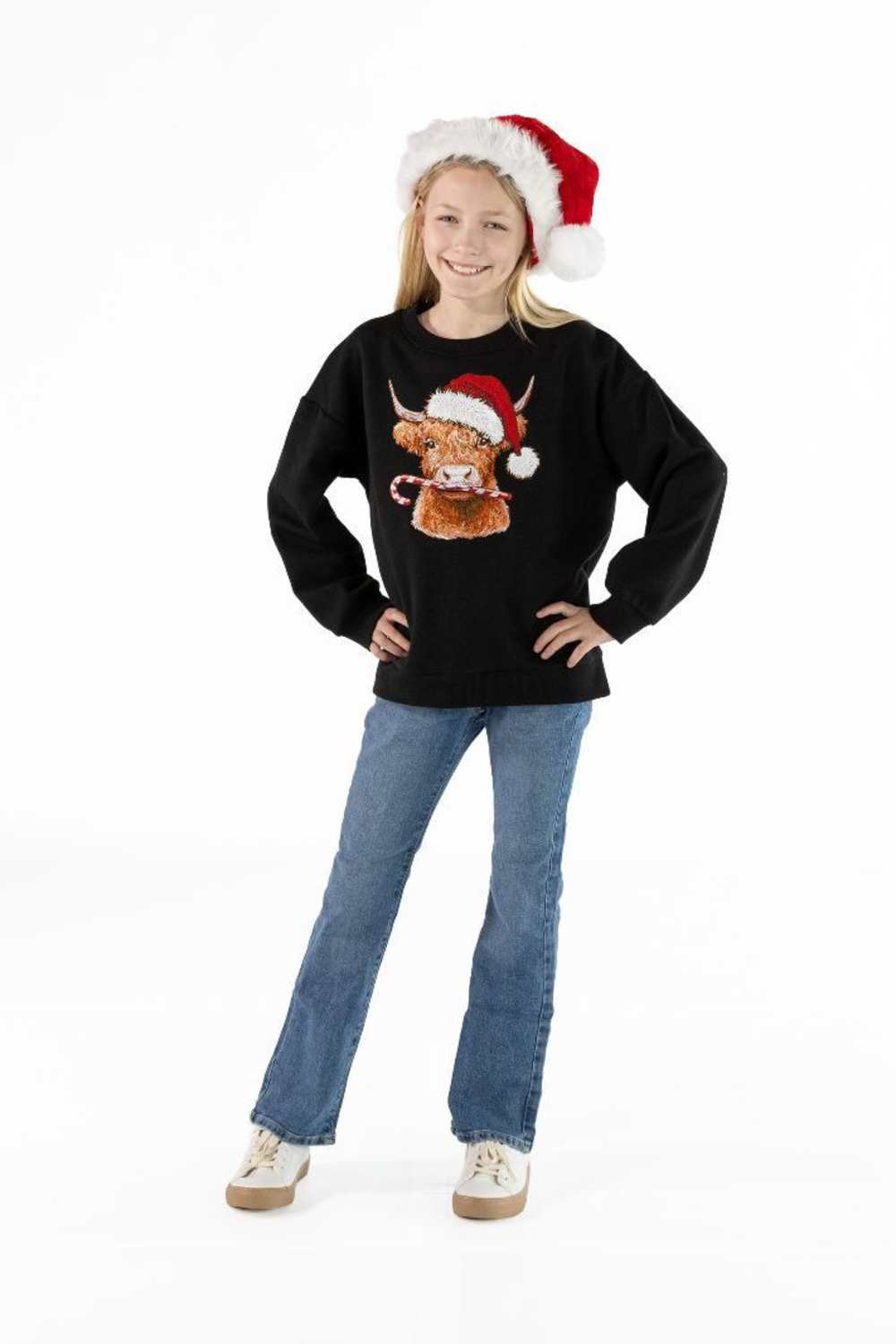 A young girl modeling a black long-sleeve crewneck sweatshirt with a graphic of a Scottish Highland cow in a Santa hat, paired with blue jeans and a matching Santa hat, on a white studio background.