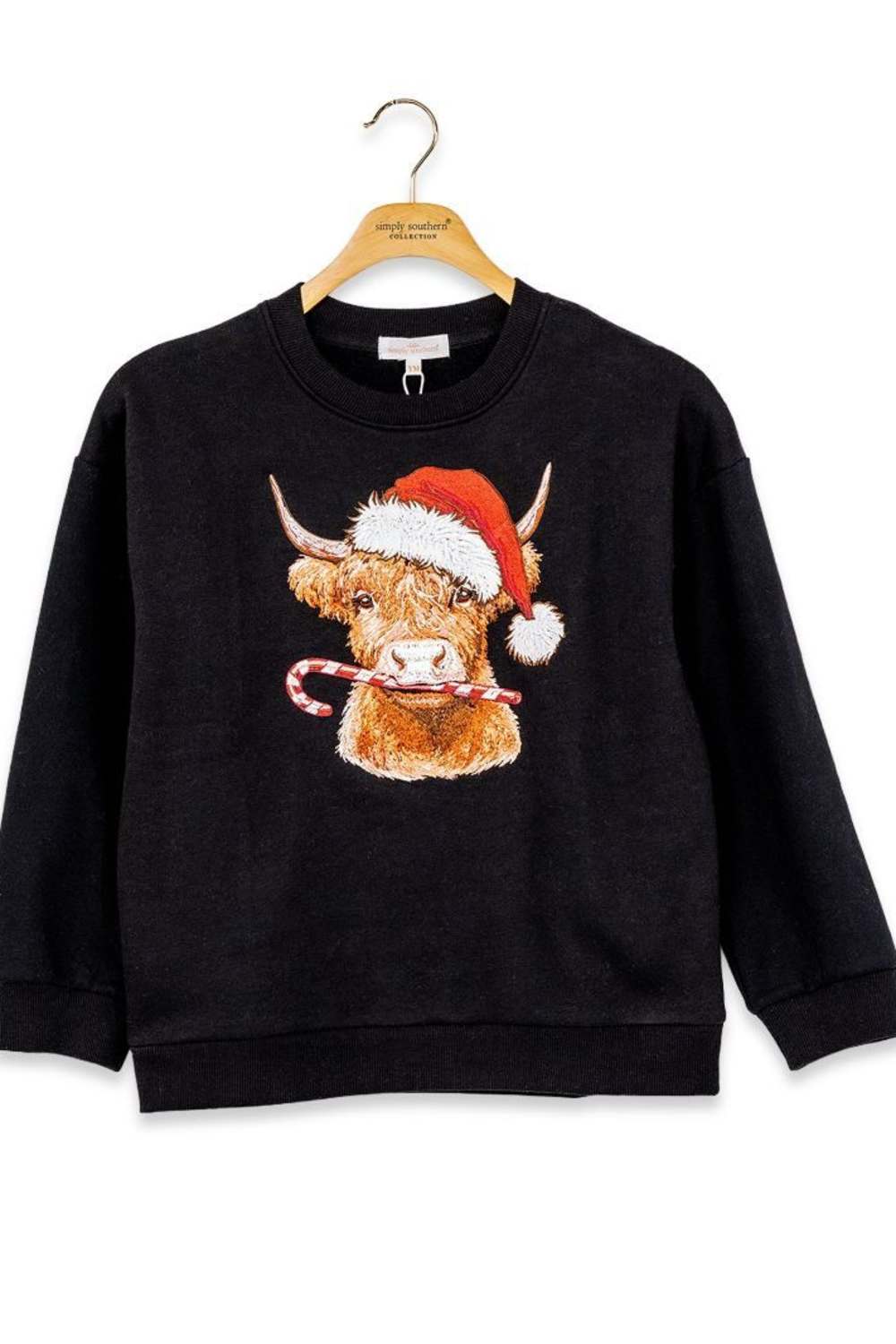 A black crewneck sweatshirt hanging on a wooden hanger, featuring a graphic of a Highland cow wearing a red Santa hat and holding a candy cane.