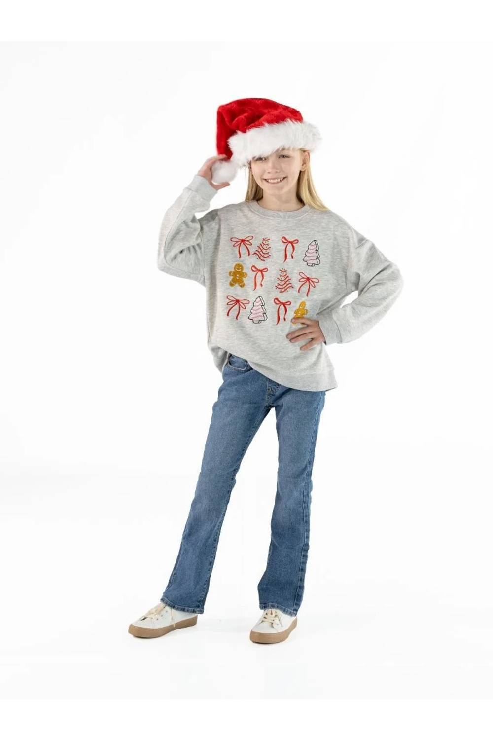 A young girl wearing a gray long-sleeve crewneck sweatshirt with a red and yellow Christmas tree pattern, blue jeans, and a red Santa hat, posing against a white studio background.