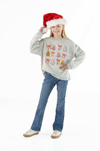 A young girl wearing a light gray long-sleeve crewneck sweatshirt with a red and yellow Christmas-themed graphic print, blue jeans, and a red Santa hat, posing against a white studio background.