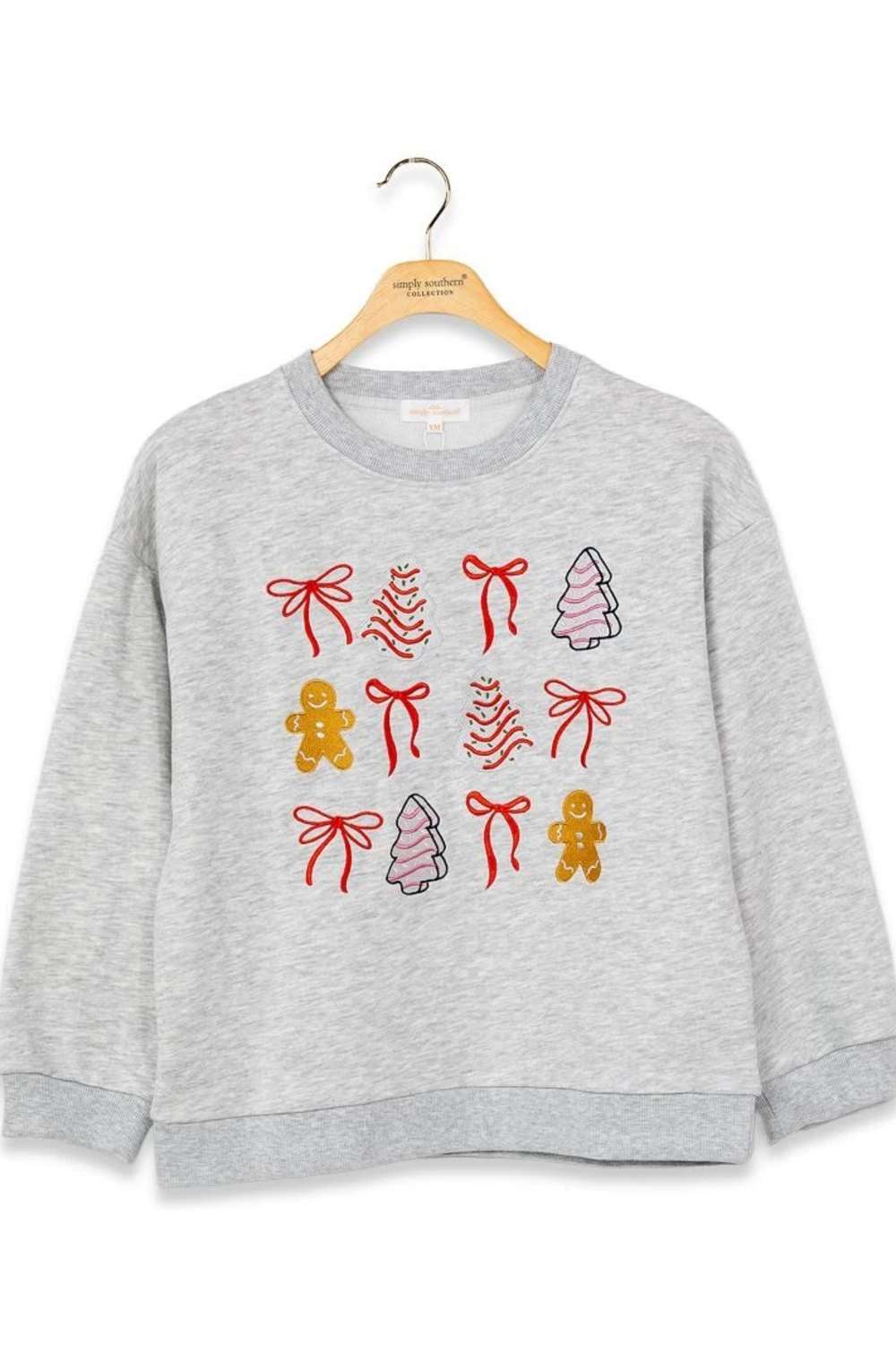 A light gray heather crewneck sweatshirt hanging on a wooden hanger, featuring a festive Christmas graphic print with red bows, gingerbread men, and decorated trees.